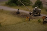 A miniature scene depicting a construction site with a small figure in a yellow jacket performing surveying work. Nearby is a trailer-mounted piece of machinery on a dirt road, with blurred greenery in the background.