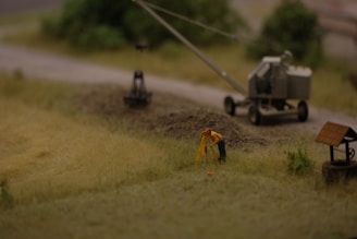 A miniature scene depicting a construction site with a small figure in a yellow jacket performing surveying work. Nearby is a trailer-mounted piece of machinery on a dirt road, with blurred greenery in the background.