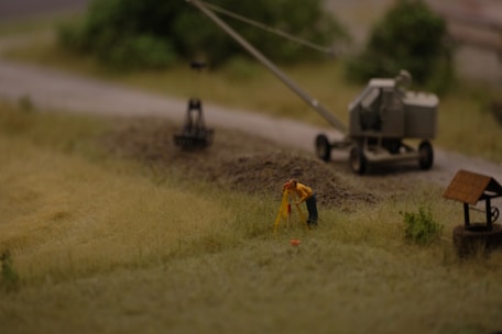 A miniature scene depicting a construction site with a small figure in a yellow jacket performing surveying work. Nearby is a trailer-mounted piece of machinery on a dirt road, with blurred greenery in the background.