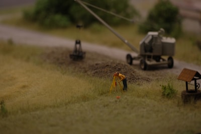 A miniature scene depicting a construction site with a small figure in a yellow jacket performing surveying work. Nearby is a trailer-mounted piece of machinery on a dirt road, with blurred greenery in the background.