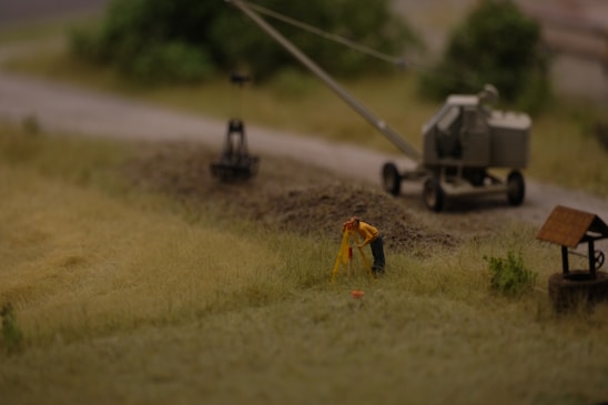 A miniature scene depicting a construction site with a small figure in a yellow jacket performing surveying work. Nearby is a trailer-mounted piece of machinery on a dirt road, with blurred greenery in the background.