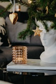 A decorative scene featuring a close-up of a Christmas tree branch with a heart-shaped cookie ornament and a star ornament. A white sculptural bust sits beside a textured amber glass candle holder on top of two magazines.