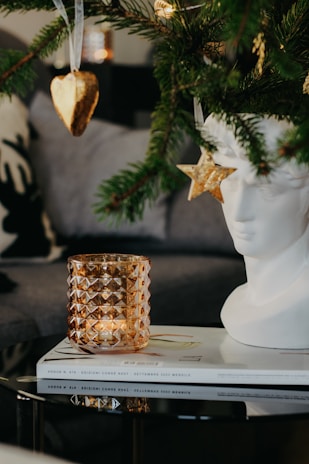 A decorative scene featuring a close-up of a Christmas tree branch with a heart-shaped cookie ornament and a star ornament. A white sculptural bust sits beside a textured amber glass candle holder on top of two magazines.