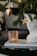 A decorative scene featuring a close-up of a Christmas tree branch with a heart-shaped cookie ornament and a star ornament. A white sculptural bust sits beside a textured amber glass candle holder on top of two magazines.