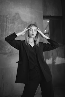 A woman in a black suit stands in front of a textured wall with a serious expression. She has long, straight hair and holds her hands to the sides of her head. The background features a slightly blurred concrete-like surface with some shading.