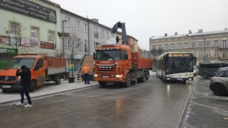 A street scene in an urban area featuring a large orange Scania truck and a bus labeled with the route number 40. There are several buildings lining the street, adorned with signs and advertisements. Two people are present; one male figure dressed in dark clothing captures a photo, and two workers in orange vests attend to the truck and a nearby van. The sky is overcast, suggesting a cold and possibly snowy day with traces of snow on the ground.