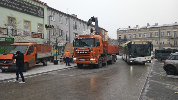 A street scene in an urban area featuring a large orange Scania truck and a bus labeled with the route number 40. There are several buildings lining the street, adorned with signs and advertisements. Two people are present; one male figure dressed in dark clothing captures a photo, and two workers in orange vests attend to the truck and a nearby van. The sky is overcast, suggesting a cold and possibly snowy day with traces of snow on the ground.