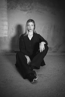 A woman with long hair is sitting on a textured floor in a relaxed pose, wearing a dark suit. The background features a roughly textured wall with varying shades and patterns.