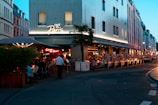 A bustling evening scene at a Parisian café with outdoor seating. People are dining under a large umbrella, alongside potted plants. A neon sign illuminates the entrance, while the building behind is modern with large windows. The street is adorned with more buildings and lit by street lamps as twilight begins.