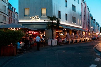 A bustling evening scene at a Parisian café with outdoor seating. People are dining under a large umbrella, alongside potted plants. A neon sign illuminates the entrance, while the building behind is modern with large windows. The street is adorned with more buildings and lit by street lamps as twilight begins.