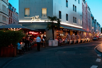 A bustling evening scene at a Parisian café with outdoor seating. People are dining under a large umbrella, alongside potted plants. A neon sign illuminates the entrance, while the building behind is modern with large windows. The street is adorned with more buildings and lit by street lamps as twilight begins.