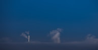 A single white smokestack emitting smoke into a dark blue sky. Clouds are visible near and around the smokestack, blending with the smoke. The scene is minimalistic with a strong contrast between the white smokestack and the deep blue background.