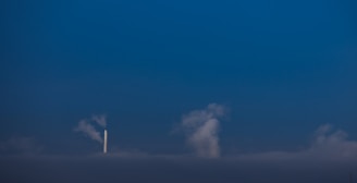 A single white smokestack emitting smoke into a dark blue sky. Clouds are visible near and around the smokestack, blending with the smoke. The scene is minimalistic with a strong contrast between the white smokestack and the deep blue background.