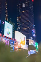 A vibrant urban scene featuring numerous illuminated billboards against the backdrop of tall buildings at night. The display screens show various advertisements, including a large image of a person in a yellow outfit. Neon lights and reflections add a lively atmosphere.