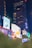 A vibrant urban scene featuring numerous illuminated billboards against the backdrop of tall buildings at night. The display screens show various advertisements, including a large image of a person in a yellow outfit. Neon lights and reflections add a lively atmosphere.