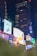 A vibrant urban scene featuring numerous illuminated billboards against the backdrop of tall buildings at night. The display screens show various advertisements, including a large image of a person in a yellow outfit. Neon lights and reflections add a lively atmosphere.