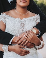 A close-up of a couple with intertwined arms, highlighting their wedding rings. The bride is wearing an intricate lace gown, and the groom is dressed in a black suit. The setting appears to be outdoors, possibly in a garden.