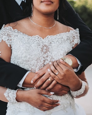 A close-up of a couple with intertwined arms, highlighting their wedding rings. The bride is wearing an intricate lace gown, and the groom is dressed in a black suit. The setting appears to be outdoors, possibly in a garden.