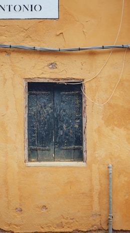 A weathered, dark wooden window is set within a rough, mustard-colored stucco wall. Above the window, there is a portion of a white sign visible with the letters 'NTONIO'. Black wires run horizontally across the wall, with one wire curving downward, and a metal pipe extends vertically from the bottom right corner of the image.