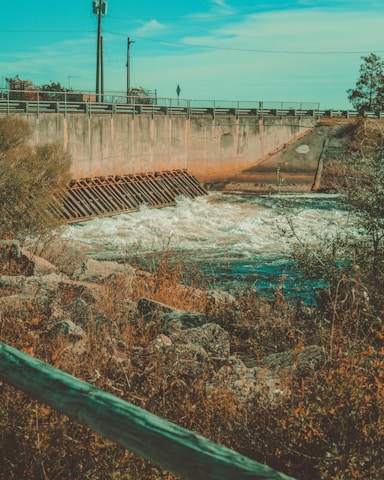 A concrete structure with a metal grate is observed holding back water, creating turbulent flow beneath it. The area is surrounded by rocky terrain with overgrown vegetation and a wooden fence in the foreground. There are utility poles and a clear blue sky in the background.