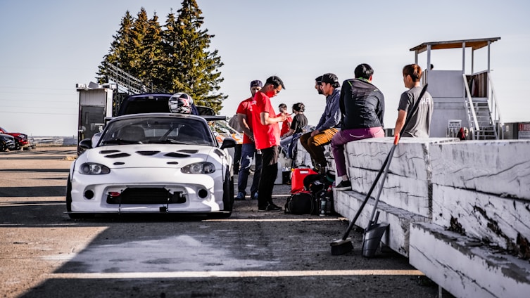 A group of people are gathered around a modified white sports car at a racing track. Some individuals are sitting on a white barrier, while others stand nearby, suggesting they may be preparing for a race. Helmets and equipment are visible, and a watchtower stands in the background.