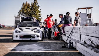 A group of people are gathered around a modified white sports car at a racing track. Some individuals are sitting on a white barrier, while others stand nearby, suggesting they may be preparing for a race. Helmets and equipment are visible, and a watchtower stands in the background.