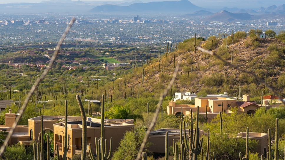 A scenic view of a desert landscape featuring numerous cacti scattered across the rolling hills. In the foreground, several adobe-style houses with flat roofs and warm-toned walls blend into the surroundings. The background reveals a sprawling cityscape with buildings rising against a backdrop of distant mountain ranges under a hazy sky.