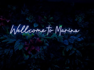 Neon blue text reads 'Wellcome to Marina' against a backdrop of lush, dark green foliage with hints of red and purple from various plants.