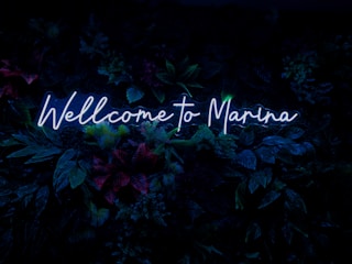 Neon blue text reads 'Wellcome to Marina' against a backdrop of lush, dark green foliage with hints of red and purple from various plants.