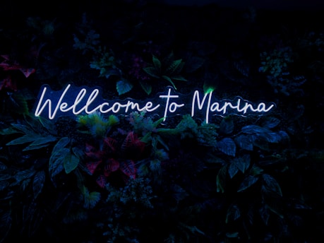 Neon blue text reads 'Wellcome to Marina' against a backdrop of lush, dark green foliage with hints of red and purple from various plants.