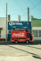 A small red Coca-Cola delivery truck is parked in front of a weathered blue storefront with a large Corona sign. The scene depicts an urban street with overhead power lines casting shadows on the ground.