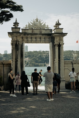 A group of people stand in front of an ornate stone archway, with a view of a large body of water and a cityscape in the background. The scene suggests a tourist attraction or historical site.