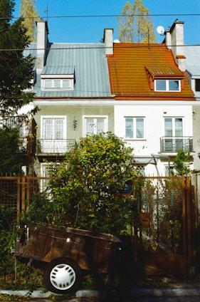 A row of two-story residential homes with distinct colored roofs, one in a muted grey and the other in a rusty red. Each house has a small balcony on the second floor, while the first floor features large windows. A lush garden with dense greenery is in front of the houses, and an old wooden cart with a white wheel is next to a brown metal fence. Tall trees are visible in the background under a clear blue sky.