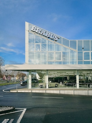 A modern building with large glass windows displays several luxury cars inside. The sign on the top left corner features the name 'BRABUS'. The sky is clear, enhancing the visibility of the sleek building design. There are trees and a pathway visible on the left side of the image.