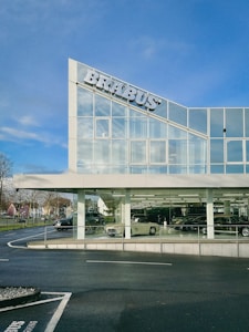 A modern building with large glass windows displays several luxury cars inside. The sign on the top left corner features the name 'BRABUS'. The sky is clear, enhancing the visibility of the sleek building design. There are trees and a pathway visible on the left side of the image.