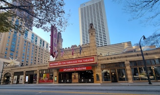 A historic theater building with intricate brickwork architecture and towers, featuring a red marquee with white lettering that reads Welcome to the Fabulous Fox Theatre, Atlanta's Theatre. Surrounding the theater are tall modern buildings, and a street with streetlights and trees is visible.