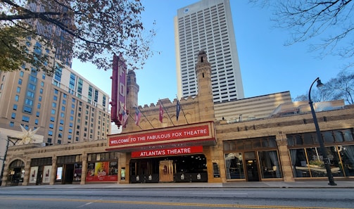 A historic theater building with intricate brickwork architecture and towers, featuring a red marquee with white lettering that reads Welcome to the Fabulous Fox Theatre, Atlanta's Theatre. Surrounding the theater are tall modern buildings, and a street with streetlights and trees is visible.