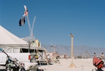 A large tent with a vibrant flag above it stands in a sandy, open area. Bicycles and a white truck are visible in the foreground. A wooden pole is in the center, with mountains in the distance under a clear blue sky.