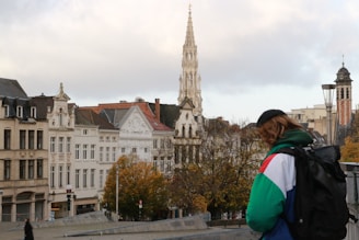 A cityscape with historical buildings featuring ornate architecture, including a tall spire. Autumn trees with golden leaves are visible, and a person wearing a colorful jacket and backpack is in the foreground.