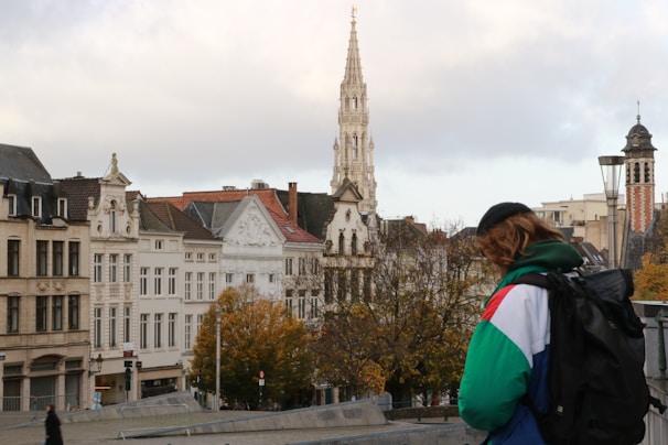 A cityscape with historical buildings featuring ornate architecture, including a tall spire. Autumn trees with golden leaves are visible, and a person wearing a colorful jacket and backpack is in the foreground.