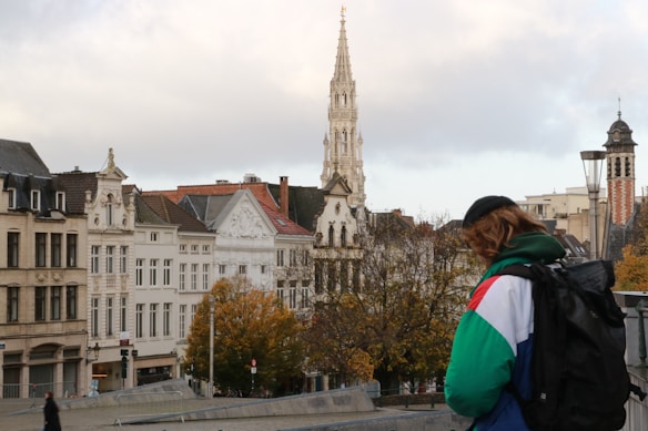A cityscape with historical buildings featuring ornate architecture, including a tall spire. Autumn trees with golden leaves are visible, and a person wearing a colorful jacket and backpack is in the foreground.