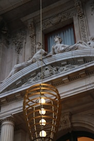 Two stone sculptures of reclining classical figures adorn an ornate architectural facade. The intricately carved details feature floral and geometric patterns. A modern, cylindrical pendant light with multiple bright bulbs hangs prominently in the foreground, creating a contrast with the historic stonework.