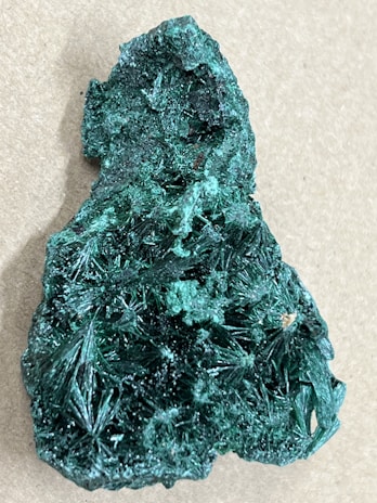 A vibrant green mineral with a rough and jagged surface, featuring intricate crystalline formations resembling needles or stars. It has a textured and shimmering appearance, set against a neutral background.