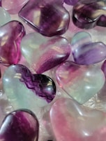 A collection of polished, heart-shaped stones with smooth surfaces. The stones are predominantly purple, pink, and translucent, with some exhibiting unique patterns and color gradients.