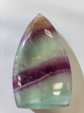 A polished, teardrop-shaped stone with a smooth surface. It features horizontal bands in shades of light purple, green, and hints of amber.