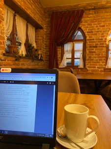 A cozy writing nook with a laptop, notebooks, and a steaming cup of tea by a sunny window.