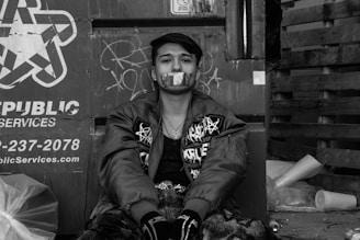 A person is sitting against a metal surface with graffiti, wearing a jacket adorned with various designs and patterns. The individual's mouth is covered with tape featuring star patterns. Surrounding the person are items like trash bags, wooden pallets, and other debris.