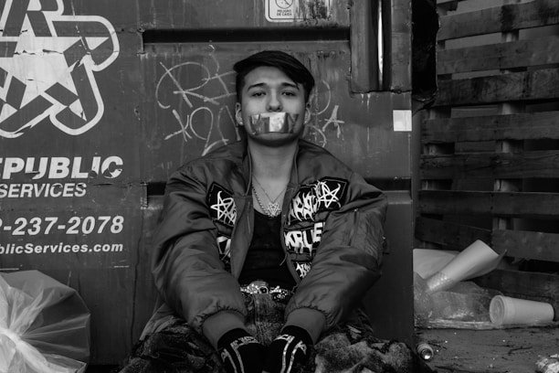 A person is sitting against a metal surface with graffiti, wearing a jacket adorned with various designs and patterns. The individual's mouth is covered with tape featuring star patterns. Surrounding the person are items like trash bags, wooden pallets, and other debris.