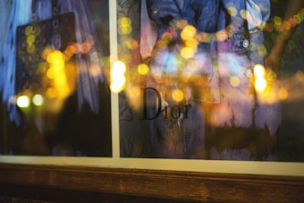 A close-up of a window display showcasing a Dior product, partially obscured by the reflection of warm, blurry lights. The scene includes hints of fashion elements such as designer clothing and a handbag, enveloped in a dreamy and artistic bokeh effect.
