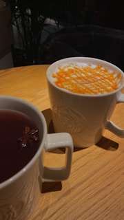 Two white ceramic mugs sit on a wooden table. One contains a cappuccino with a layer of frothy milk topped with a criss-cross pattern of orange-colored powder. The other mug holds a dark tea with a star anise floating on top. The background is slightly blurred with dark hues, possibly indicating a cafe setting.